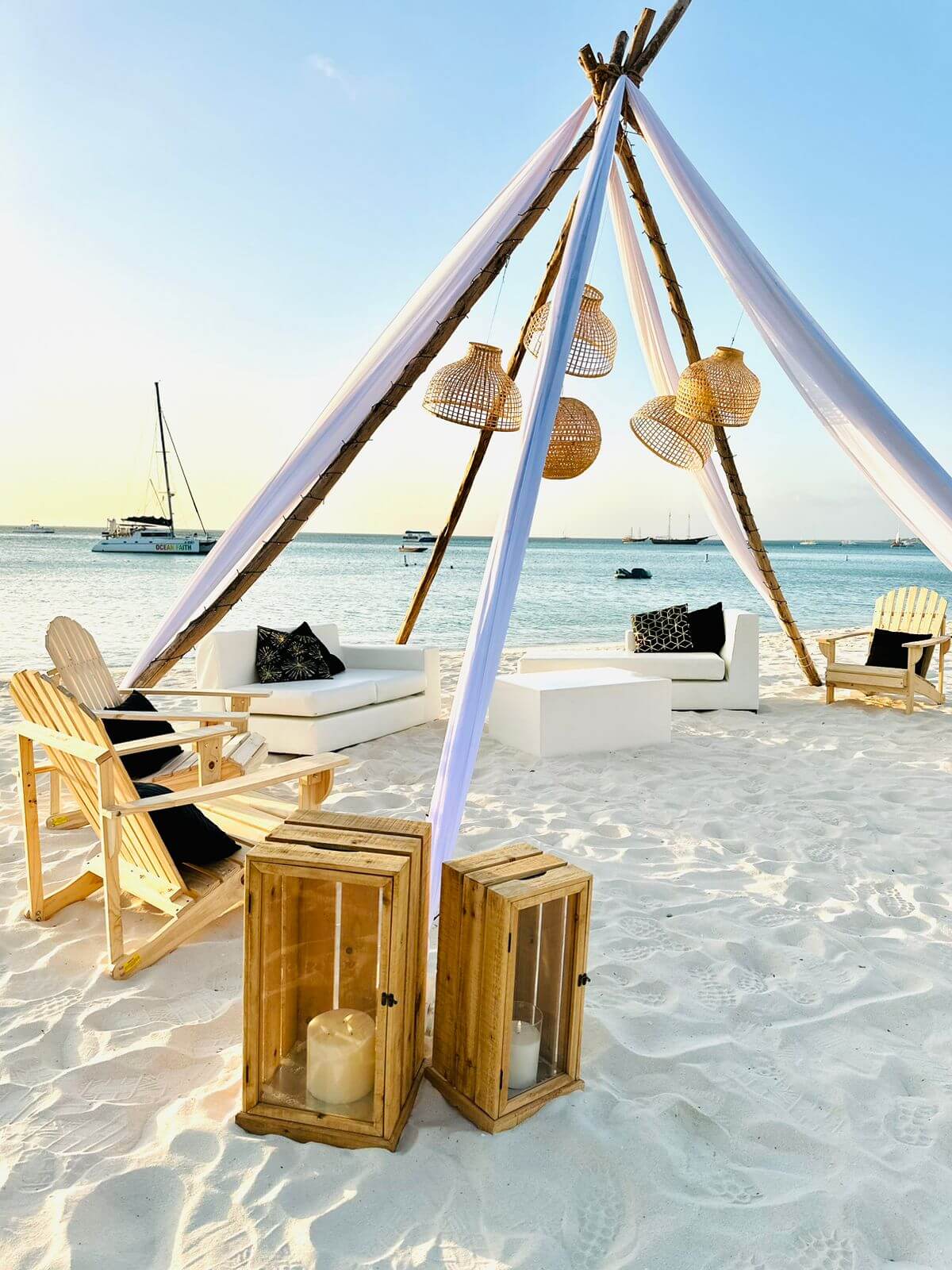 Beach canopy set up with Adirondack chairs, and couches under a structure with hanging rattan lanterns on the beach 