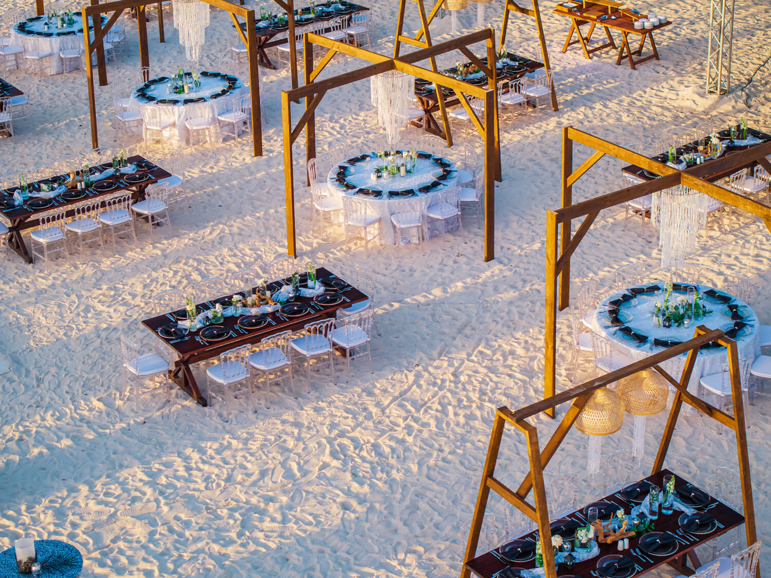 Beach event set up in the sand with tables and chairs and wooden structures holding lighting 