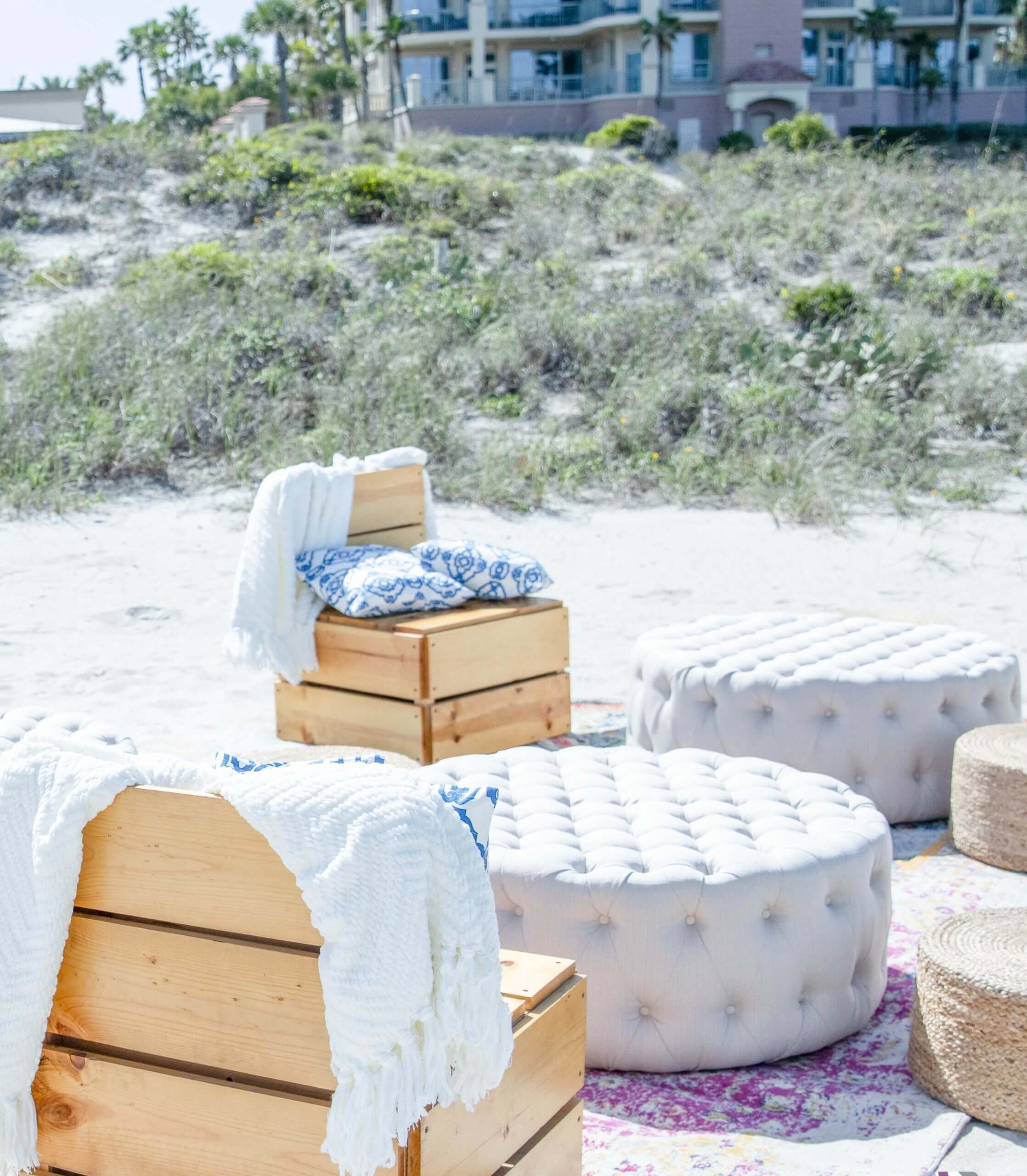 Wood and coastal whites and blue beach lounge set up with tufted ottomans on rug on sand